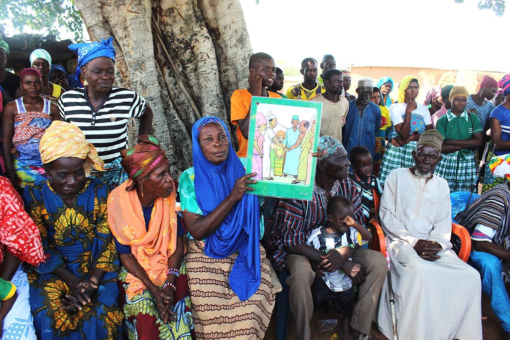 De nombreuses personnes, assises ou debout, se sont rassemblées à l'occasion d'une réunion de mobilisation communautaire à Tangbini, au Ghana. Une femme, assise au centre, tient une affiche évoquant le mariage des enfants. 